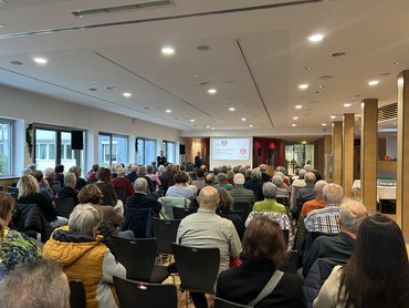 Vortrag mit vielen Personen im Saal, vorne stehen zwei Ärzte vor der Leinwand