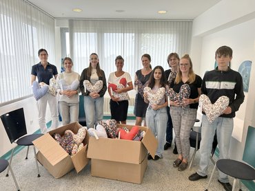 Eine Gruppe mit Menschen im Hintergrund (Schülerinnen und Schüler mit drei Lehrerinnen und einer Pflegekraft und einer Ärztin der Frauenklinik). Sie halten alle Herzkissen in der Hand. im Vordergrund stehen Kartone mit weiteren Herzkissen.